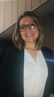 A professional portrait of a smiling woman with shoulder-length brown hair and glasses, wearing a black blazer over a white blouse.