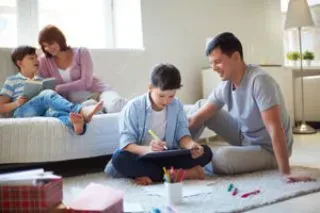 A family of four spends time together in a bright living room; two adults sit on a bed talking to a child, while another child sits on the floor drawing with colored pencils.