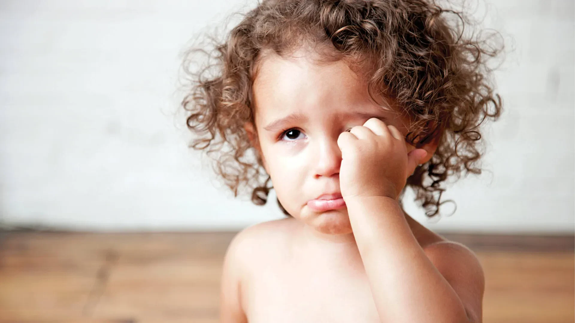 An eye-level, medium close-up shot captures a young child with curly brown hair looking sad while wiping away tears with one hand.