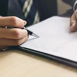 An image showing a close-up view of a person in business attire holding a pen, poised to sign or write on a blank white document placed inside a black folder on a light-colored wooden desk.