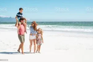 A family of four walking along a sunny white sand beach with the blue ocean waves in the background, featuring a father carrying his son on his shoulders alongside his wife and daughter.