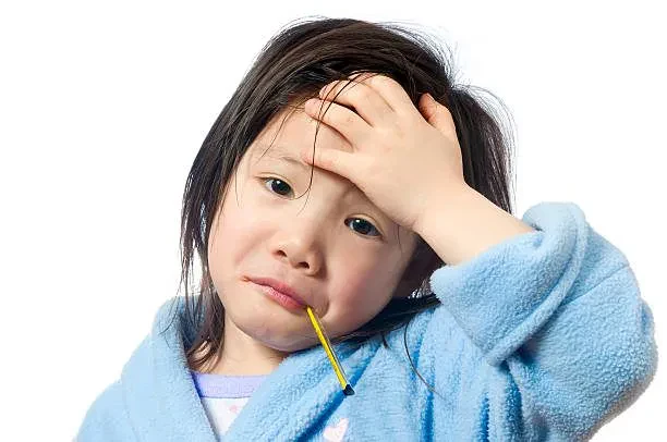 A young child with dark hair looks distressed while holding a hand to their forehead and keeping a glass thermometer in their mouth, wearing a blue fleece bathrobe against a white background.