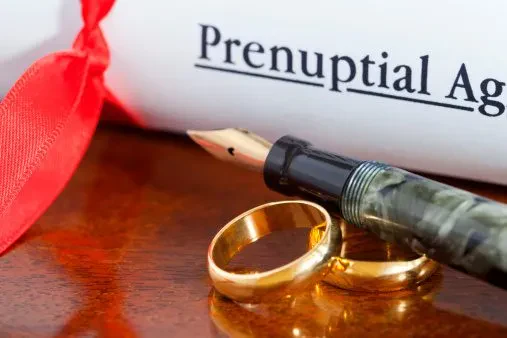 An image featuring a rolled-up document labeled "Prenuptial Agreement" tied with a red ribbon, lying next to a marble-patterned fountain pen and two gold wedding bands on a polished wooden surface.