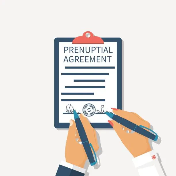 A clipboard displaying a "PRENUPTIAL AGREEMENT" document being signed by two hands, each holding a blue pen.