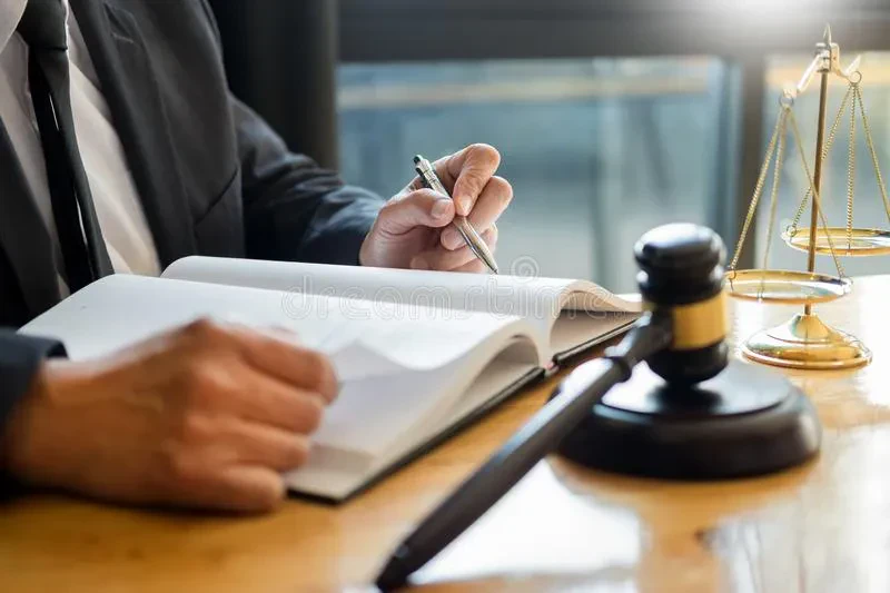 A lawyer in a black suit sits at a wooden desk, writing in a large open book with a silver pen. In the foreground, a wooden judge's gavel rests on its block, and a golden scale of justice stands to the right.