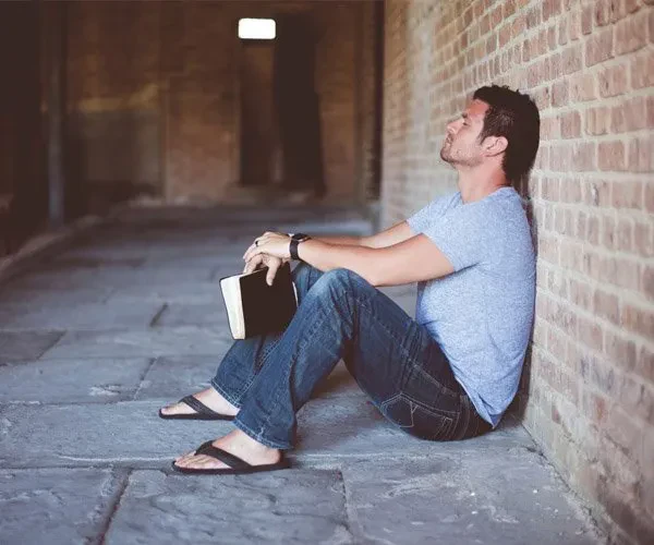 A side-view shot shows a man sitting on a stone floor with his back resting against a brick wall, his eyes closed as if in deep thought or prayer. He wears a grey t-shirt, jeans, and flip-flops, holding a book in his lap.