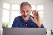 A smiling older man with grey hair waving hello during a virtual video conference call on a laptop computer inside a bright living room.