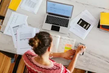 A woman seated at a table, working on a laptop with papers spread out in front of her.