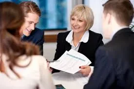 An elderly female executive smiles while presenting financial charts on documents to a group of three young colleagues during a business meeting in a modern office conference room.