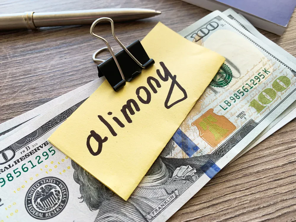 A stack of hundred-dollar bills with a yellow note labeled alimony clipped on top, placed on a wooden table.