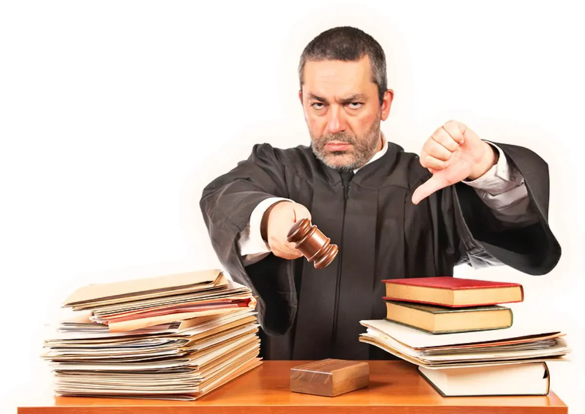 An angry judge in a black robe holding a wooden gavel out toward the camera while giving a thumbs-down gesture behind a desk stacked with law books and legal files.