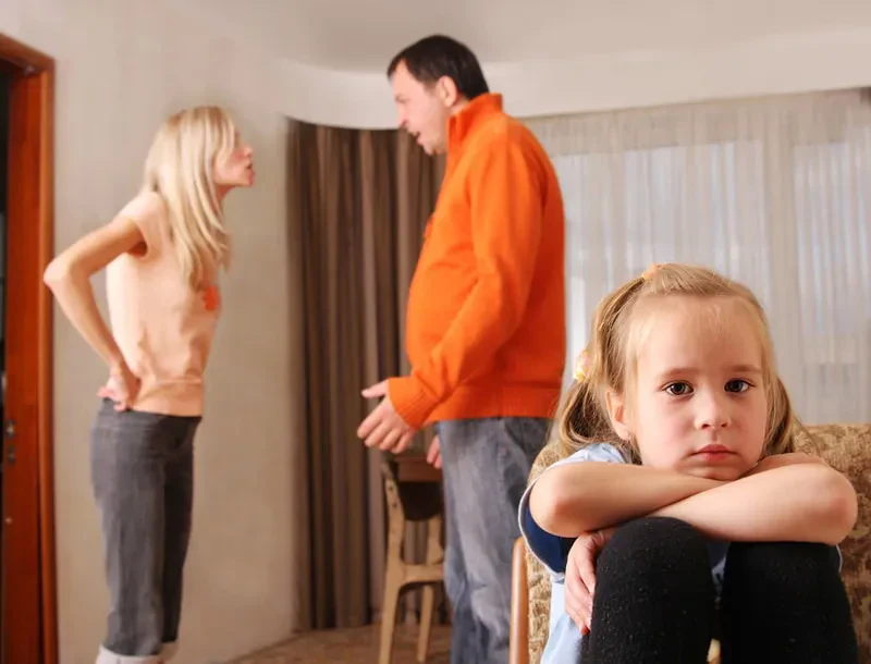 A sad young girl sits while a her parents argue in the background of a living room