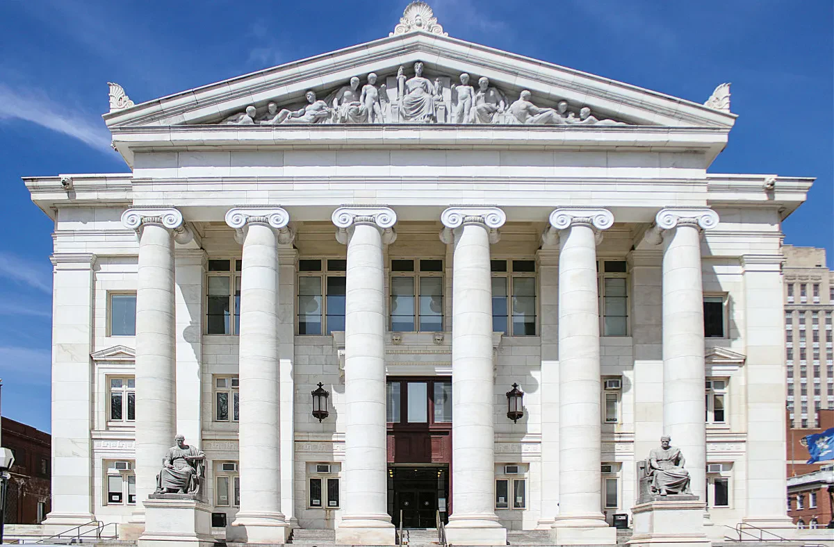 An imposing white marble building designed in the classical Beaux-Arts style, featuring six large Ionic columns supporting a triangular pediment with intricate relief sculptures.