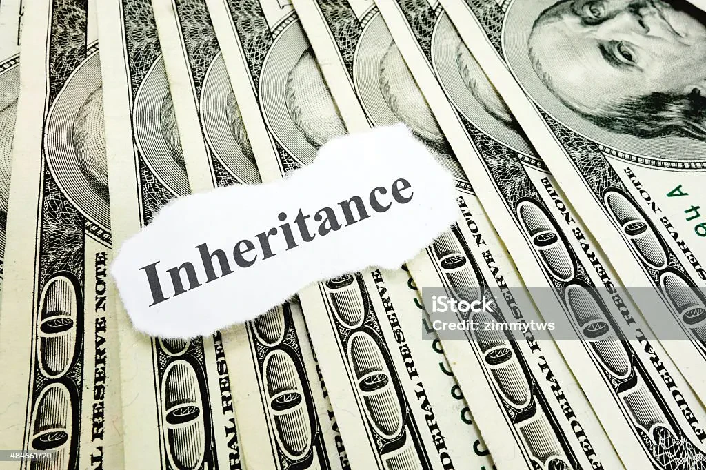 A torn piece of white paper with the word "Inheritance" printed on it lies on top of a fanned-out pile of US one hundred dollar bills.