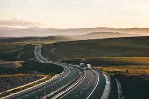 A wide highway curves through a vast, rolling landscape of golden hills and distant mountains under a hazy, warm sunset sky, with a few white trucks and cars traveling along the road.
