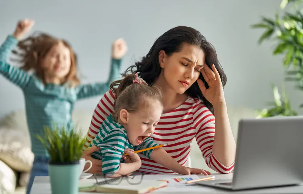 An adult woman sits at a desk with a laptop, looking stressed with one hand on her temple and her eyes closed. She is holding a small, crying toddler in a green striped shirt on her lap. In the background, another young child is jumping up and down excitedly with arms raised, creating a chaotic environment.