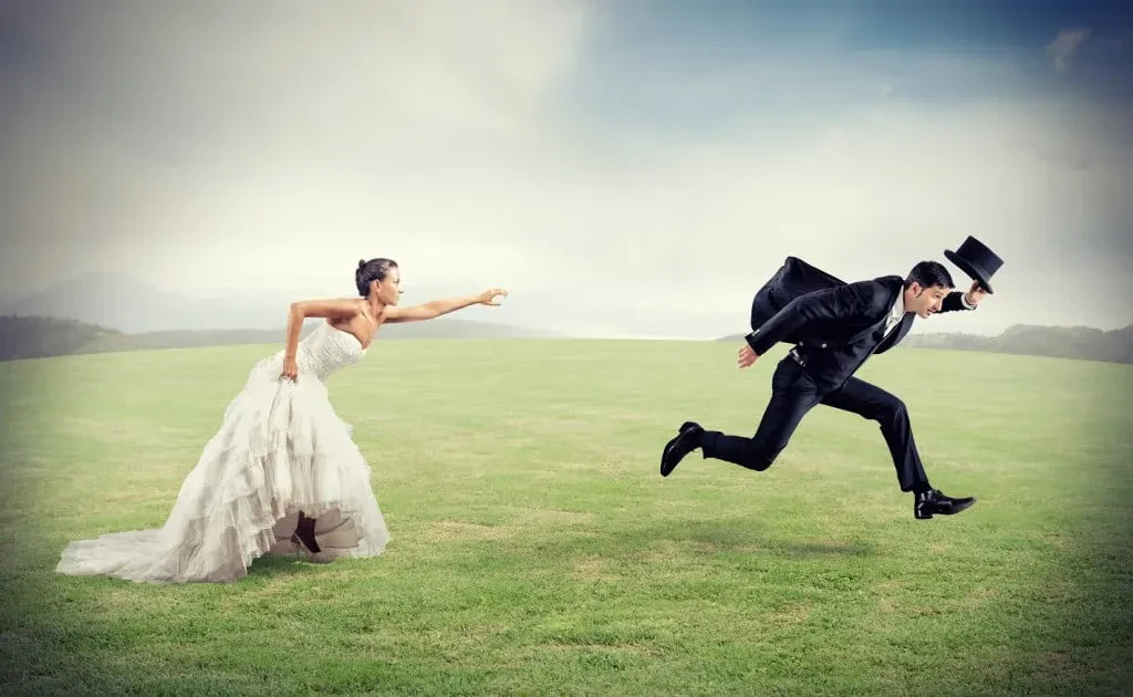 A bride in a white gown chases a groom sprinting away across a green field.