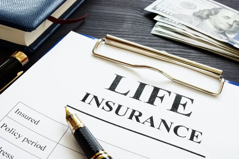 An office desk with a clipboard holding a life insurance form, featuring a fountain pen, a blue ledger, and cash.