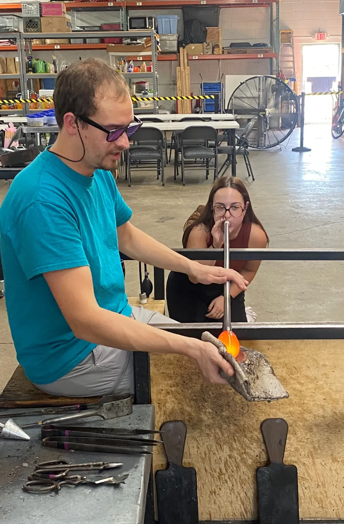 A man in a turquoise T-shirt, grey shorts, and tinted safety glasses sits at a glassblowing workbench, using a wooden block tool to shape a glowing, molten orange glass bubble on the end of a long metal blowpipe. Kneeling behind the workbench, a woman with long dark hair and glasses blows into the opposite end of the pipe to inflate the glass.
