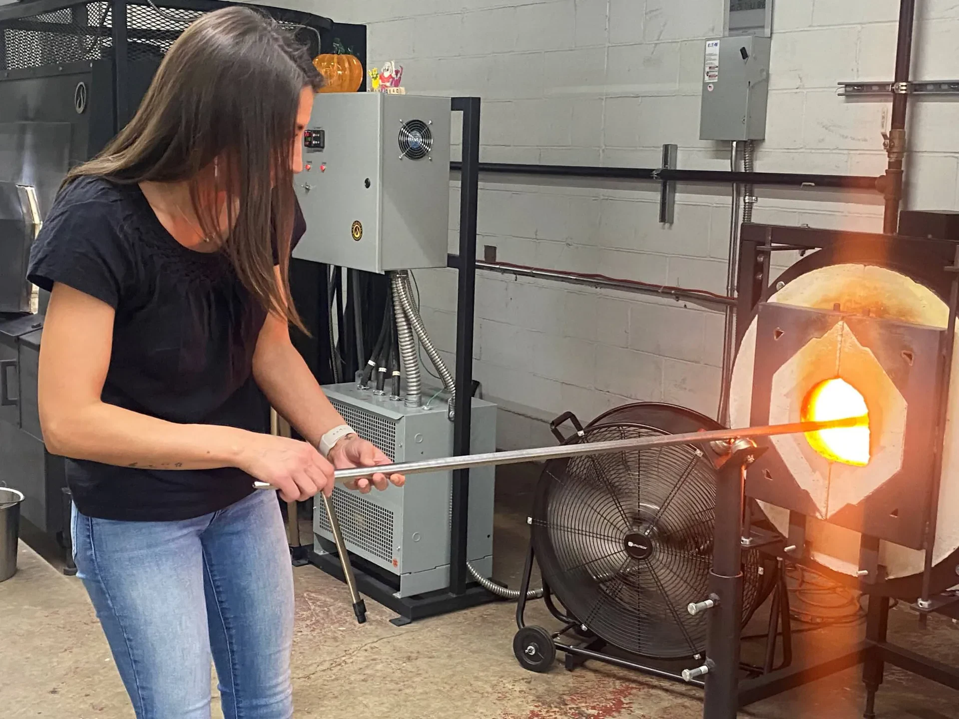 A woman with long brown hair, wearing a black short-sleeved blouse and blue jeans, stands in a workshop holding a long metal blowpipe. The rod extends into the glowing orange opening of a cylindrical glassblowing furnace.