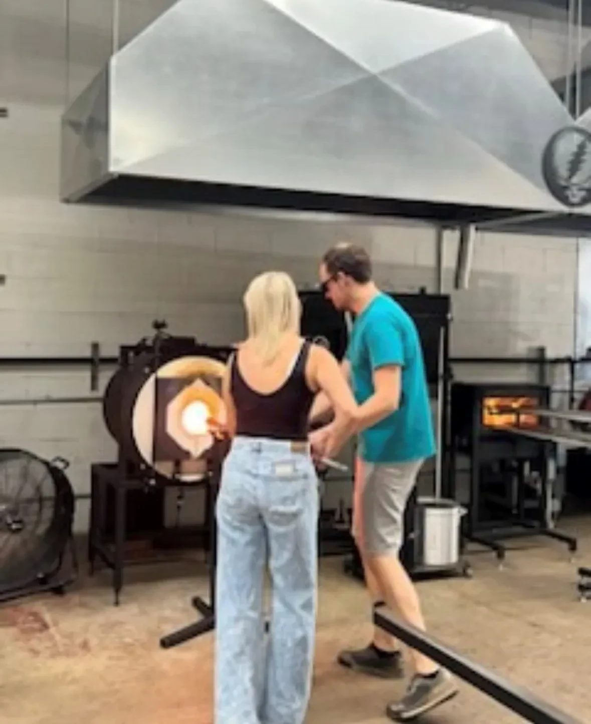 Kristen Wolf, wearing a black tank top and light blue jeans, works with a blowpipe at a glowing glassblowing furnace. A man in a turquoise T-shirt and grey shorts stands next to her, assisting.