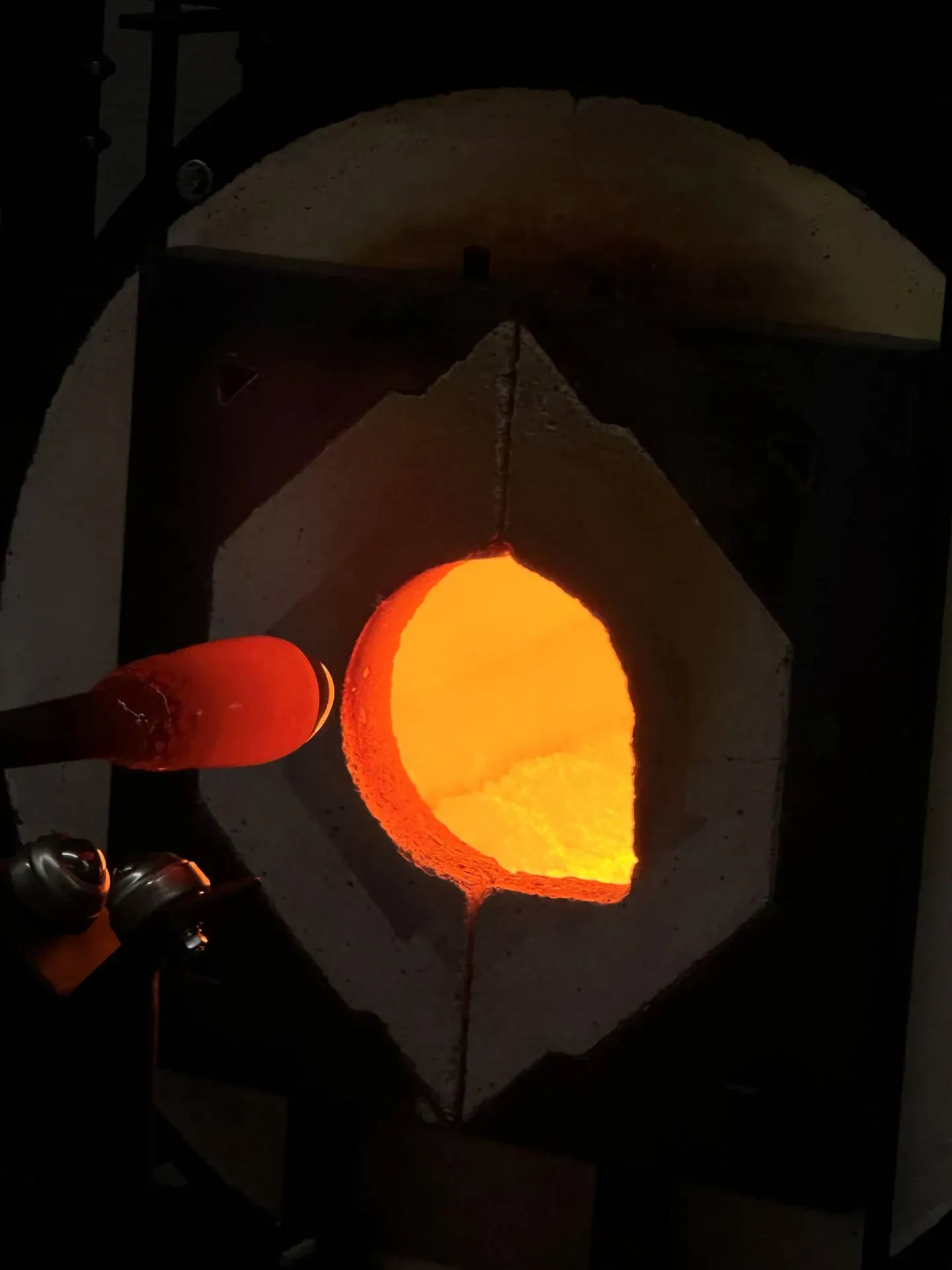 A glowing orange opening of a circular glassblowing furnace, also known as a glory hole, with a rod holding a piece of molten glass being inserted or removed.