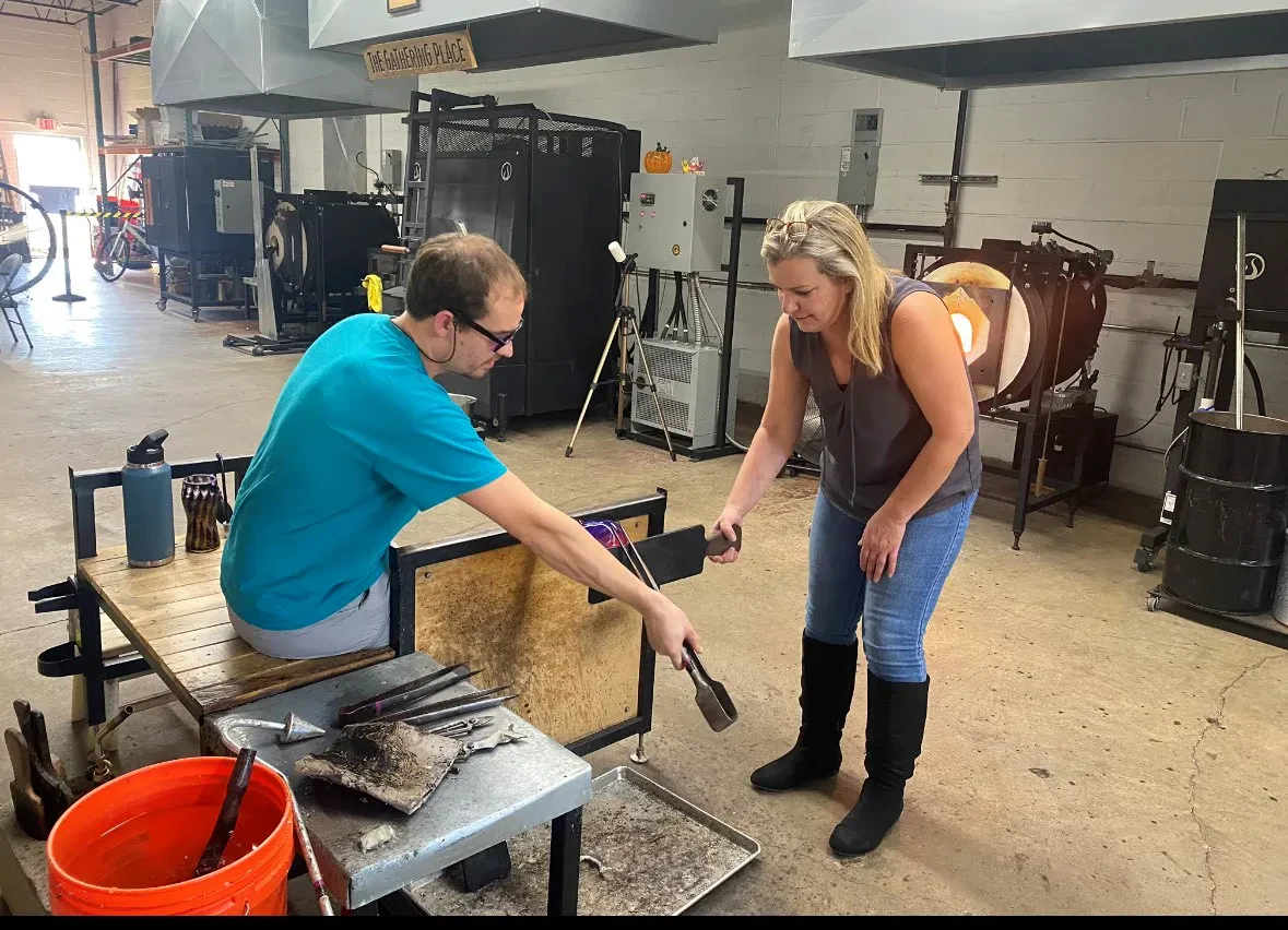 A man in a turquoise T-shirt sits at a wooden workbench shaping a glowing piece of molten glass with a hand tool. A blonde woman in a grey tank top, jeans, and black boots stands next to him, assisting and holding a tool attached to the piece.