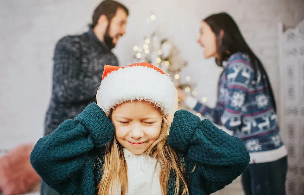 A young girl in a Santa hat shuts her ears while her parents argue