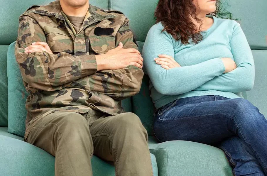 A man in military uniform and a woman in casual clothes sit on a couch with arms crossed, facing away from each other, appearing upset or in disagreement.