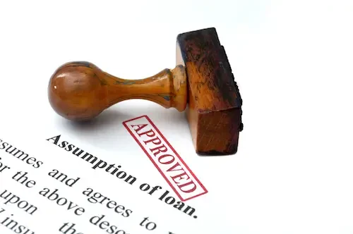 A wooden rubber stamp labeled APPROVED rests on a document titled Assumption of loan. The word APPROVED is stamped in red ink across the paper.