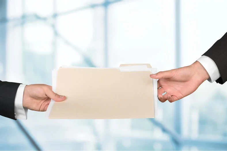 An image showing two hands in suit sleeves passing a manila file folder to each other against a blurred office background.