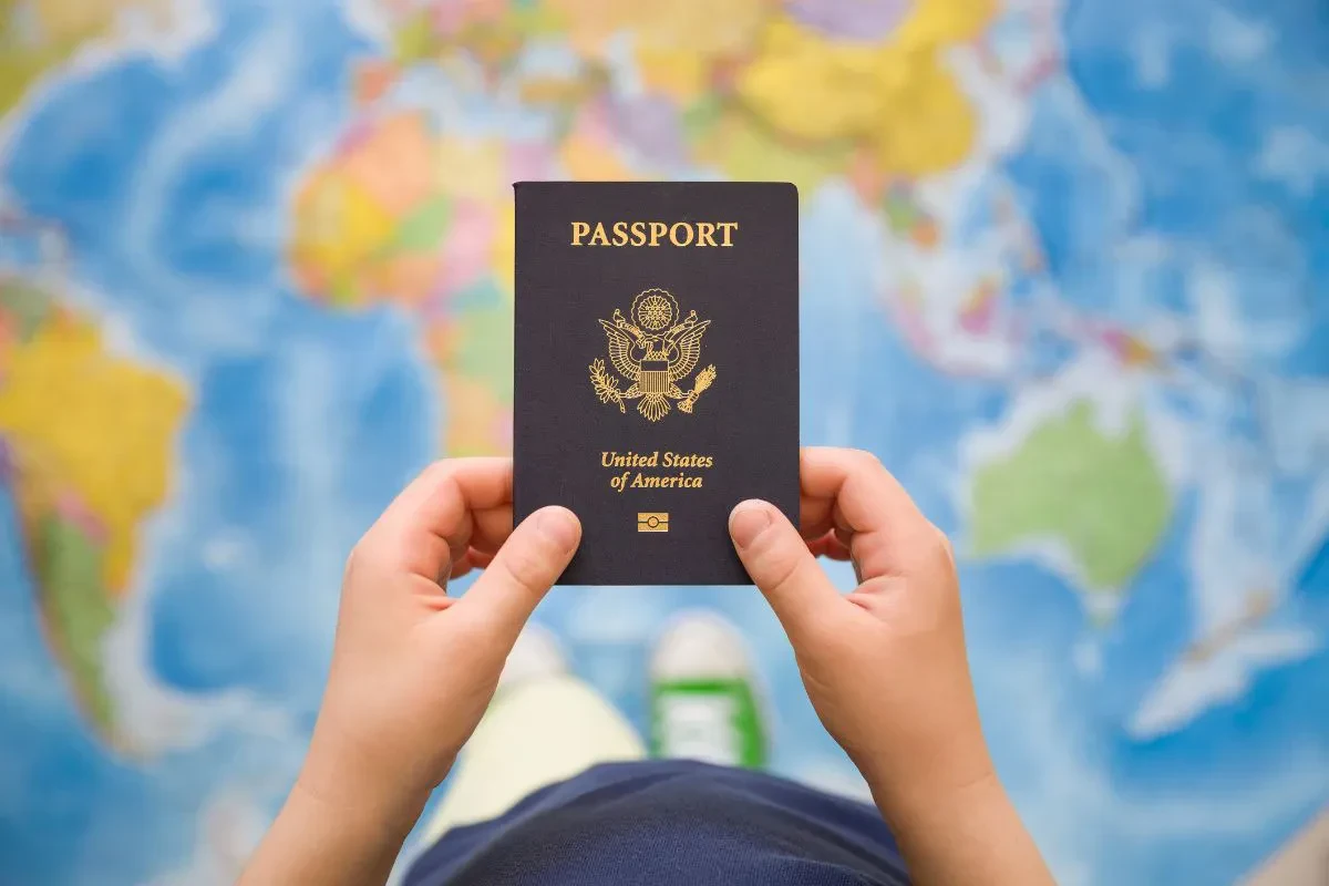 A person holds a United States passport with both hands. A colorful world map is blurred in the background.
