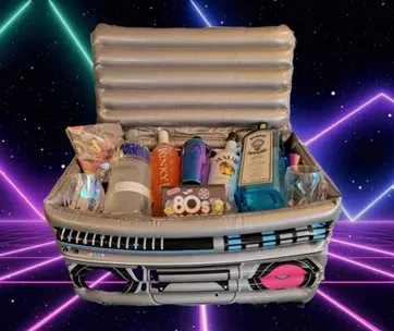 An inflatable boombox-styled cooler filled with various alcohol bottles, drinking glasses, and 1980s-themed party items against a neon retro-futuristic grid background.