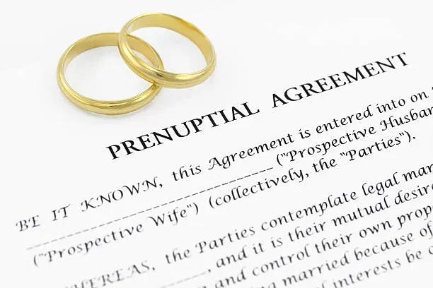 Two gold wedding rings rest on top of a prenuptial agreement document with visible text starting “PRENUPTIAL AGREEMENT.” The rings suggest a connection to marriage and legal arrangements.