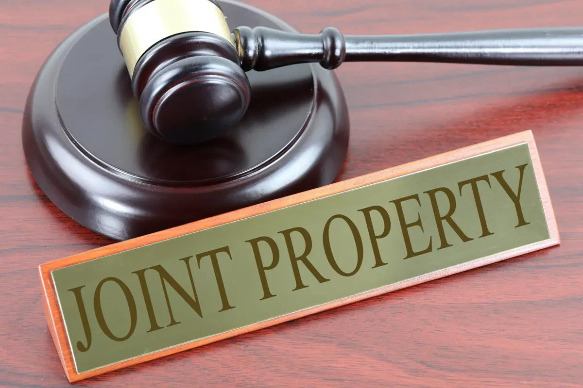 A judge’s gavel rests on a wooden table next to a nameplate that reads “JOINT PROPERTY” in bold capital letters.