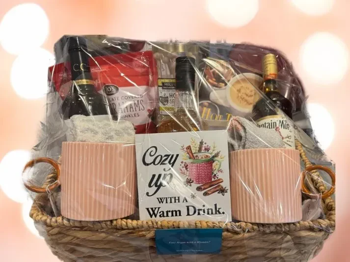 A wicker gift basket wrapped in clear cellophane, containing two pink ribbed mugs, a small sign that says "Cozy up WITH A Warm Drink," bottles of syrup or spirits, and gourmet treats.