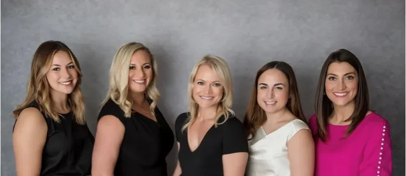 The Wolf and Shore Law Group stand side by side in front of a gray background, smiling at the camera. Four wear dark or light dresses, and one wears a bright pink top with pearl details on the sleeves.