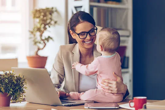 A woman wearing glasses smiles while holding a baby on her lap, working at a laptop in a home office with plants and shelves in the background.