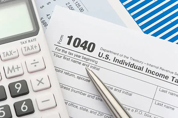 An official IRS Form 1040 U.S. Individual Income Tax Return document partially visible under a silver pen, sitting next to an office calculator and financial charts.