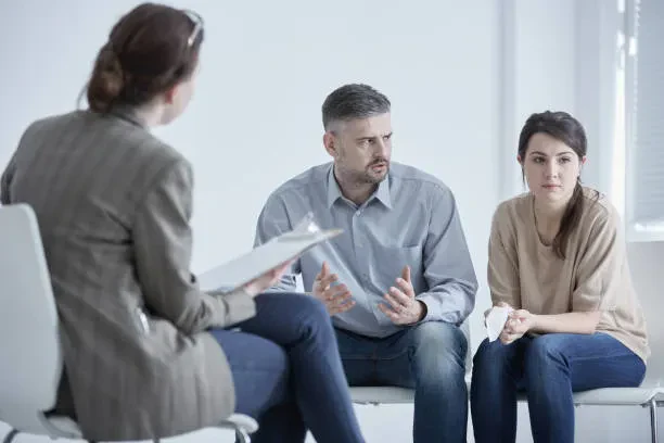 Three people sit in chairs during a counseling session; a woman with a clipboard faces a man and woman who appear to be discussing something serious.