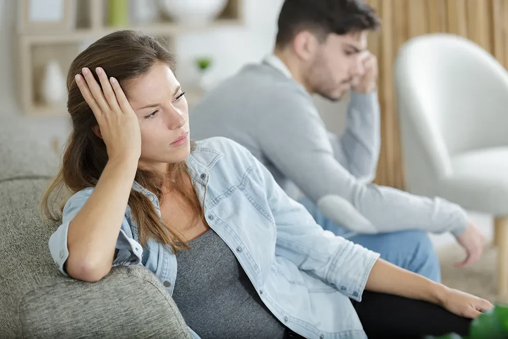 A distressed man and woman sit on a couch, each with their heads in their hands, indicating a moment of despair.
