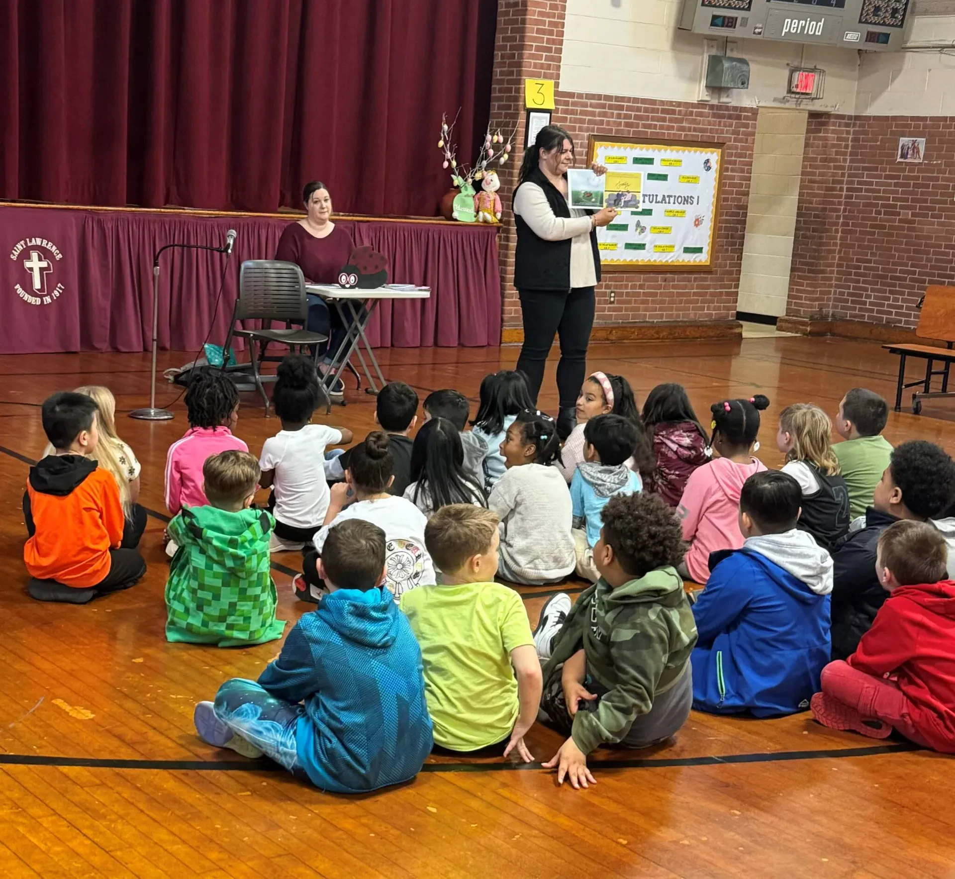 An adult woman stands on a wooden gymnasium floor, holding up a children's book to a group of about twenty young students seated in a semi-circle before her. Another woman sits in the background near a small table on a stage with a maroon curtain.