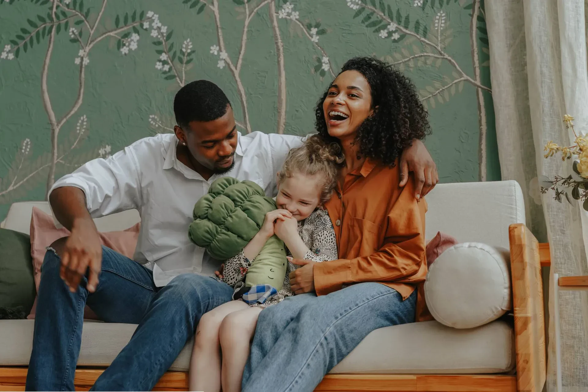 A smiling couple sits on a couch with a young girl between them, holding a plush toy. They appear happy and relaxed in a cozy, plant-themed room.
