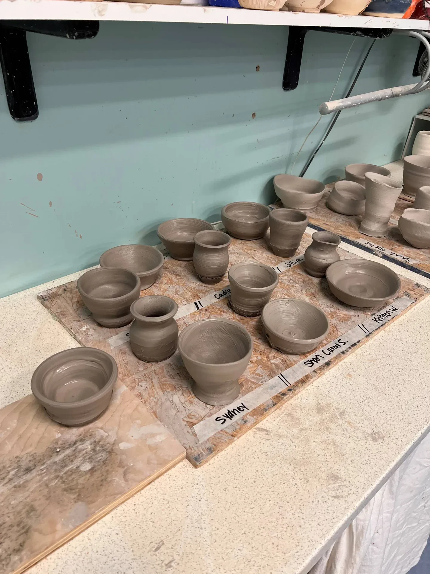 A collection of unfired, gray clay pottery pieces sitting on a wooden board inside a ceramic studio.