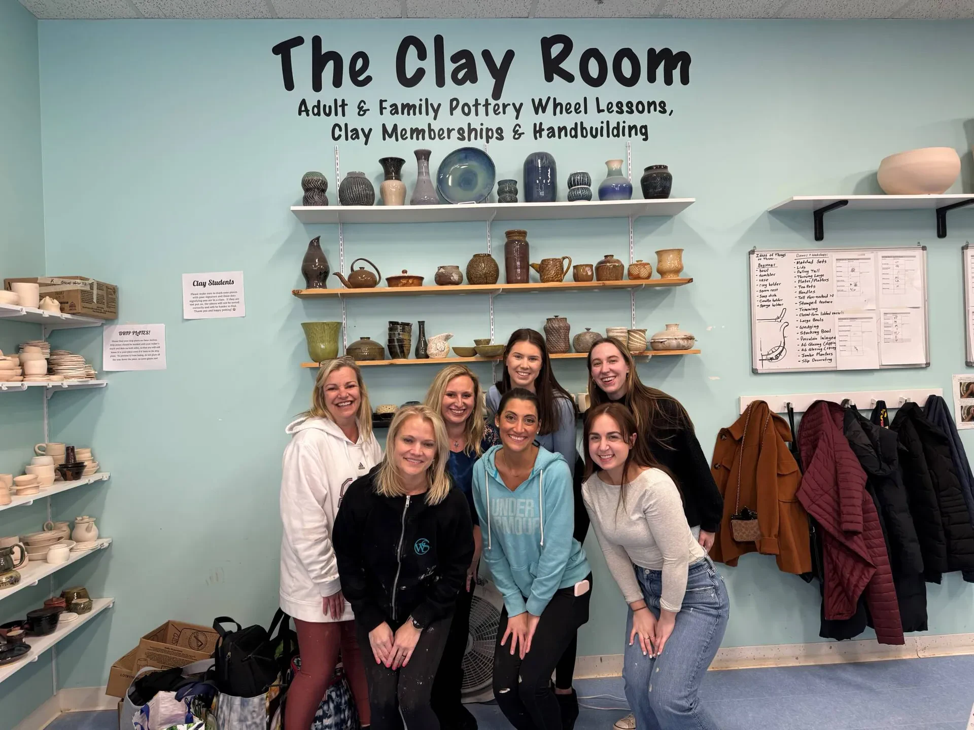 A group of women smiling and posing in front of a wall featuring a sign that reads "The Clay Room."