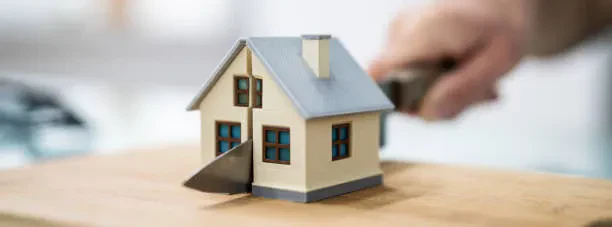 A small model house sits on a wooden cutting board, being sliced down the middle by a large kitchen knife held by an out-of-focus hand.