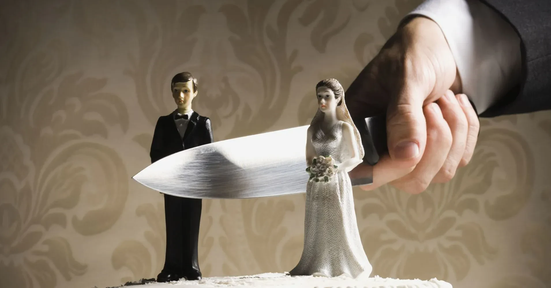 A close-up shot of a hand in a suit sleeve holding a large, sharp knife, slicing directly between a miniature bride and groom cake topper on a wedding cake against a patterned wallpaper background.