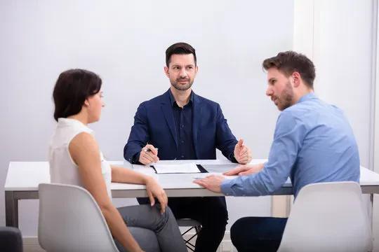 A man addresses a man and woman sitting at a table, all appearing engaged in discussion.