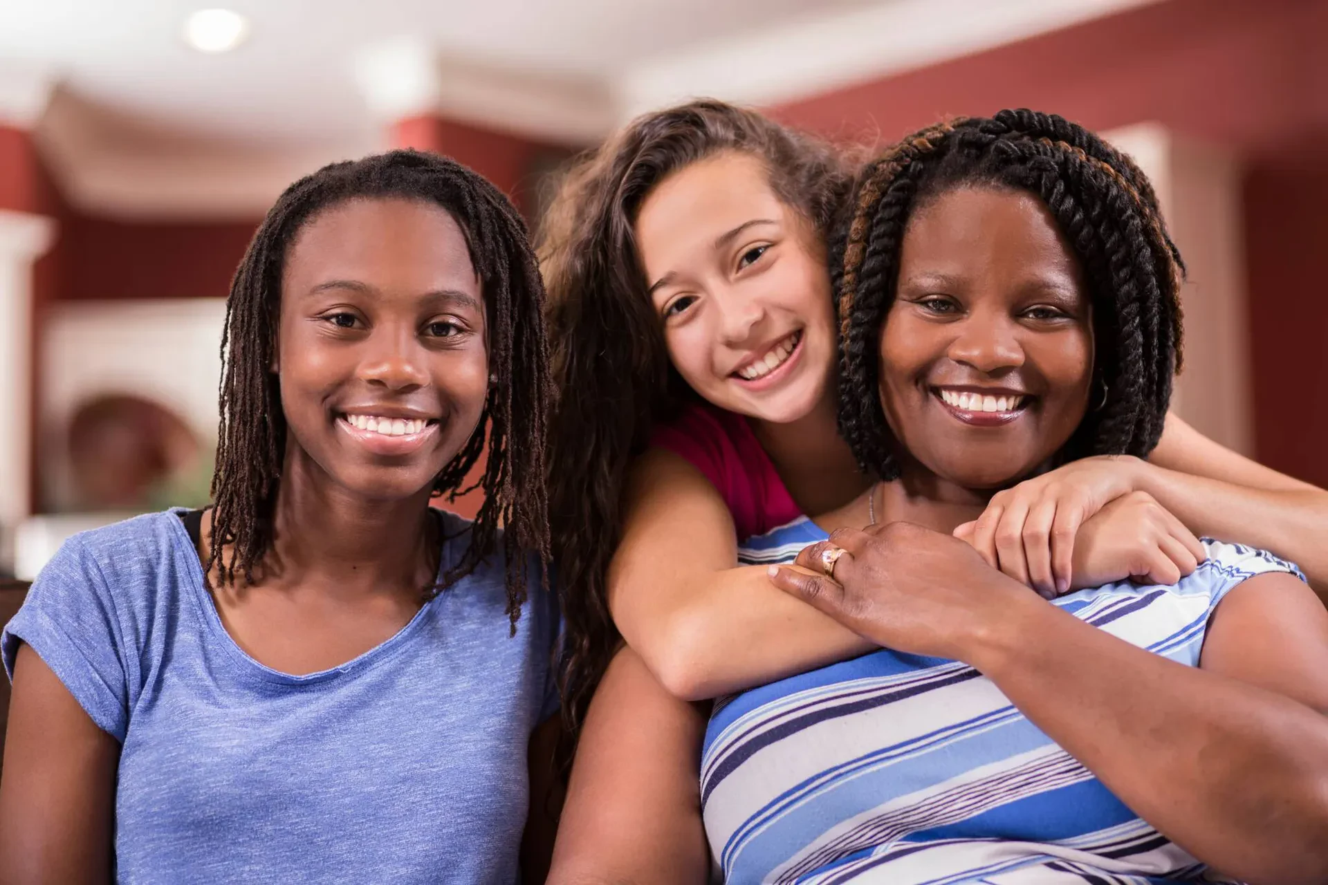 Three women are sitting close together indoors, smiling at the camera. The woman on the right is wearing a striped shirt, the center woman is hugging her from behind, and the woman on the left is wearing a blue shirt.