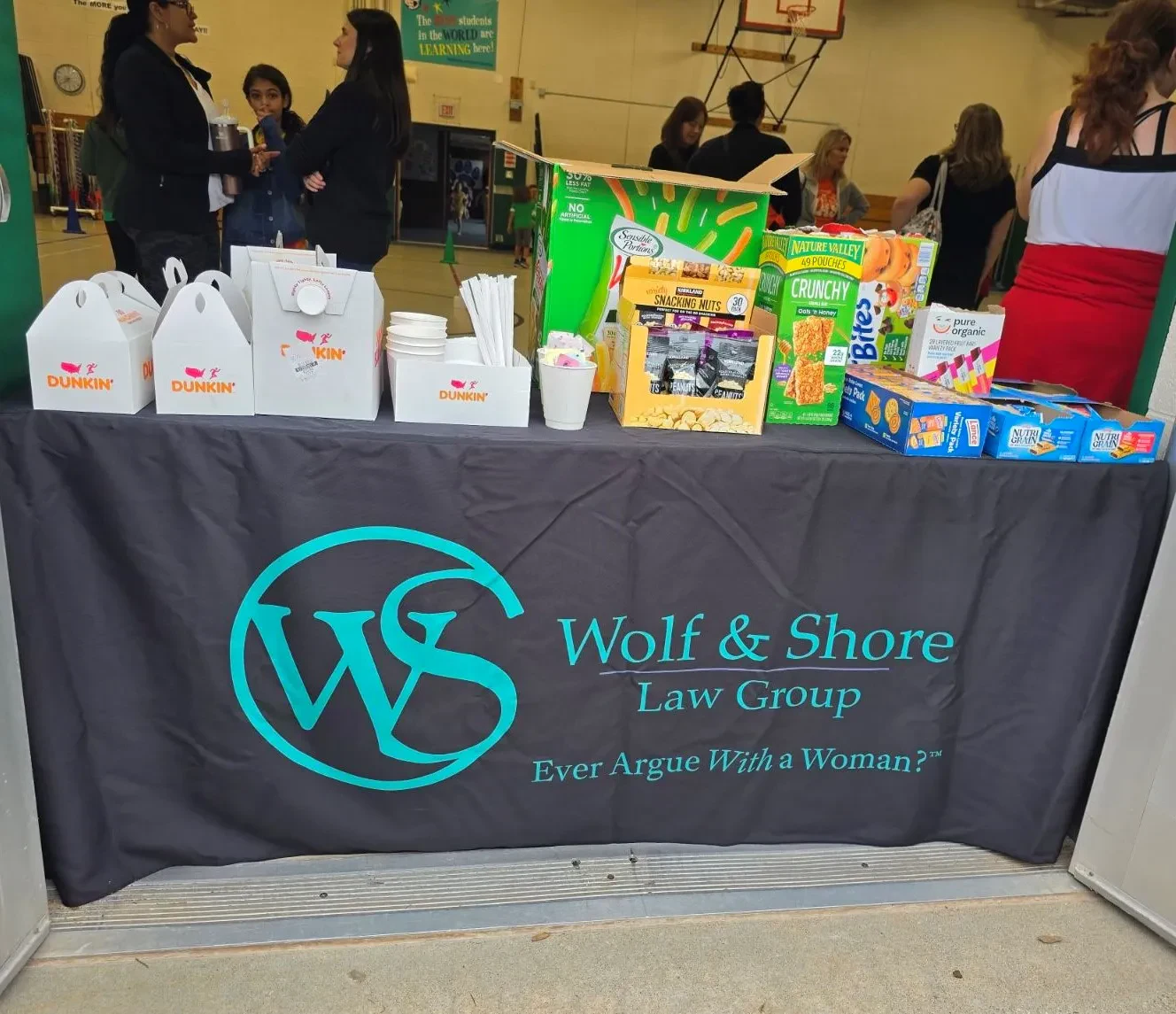 A table with snacks, Dunkin boxes, and drinks sits behind a black Wolf & Shore Law Group banner. People gather in the background at an indoor event.